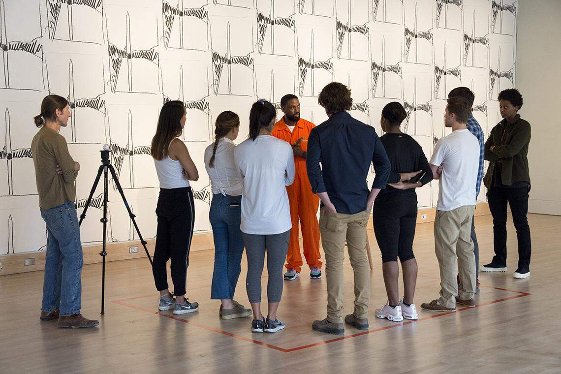 Sherrill Roland wore an orange jumpsuit for a type of performance art called the Jumpsuit Project to ignite conversations about incarceration and criminal justice. Here, he has invited the public to stand inside a cell-sized space he has created with tape on the floor.