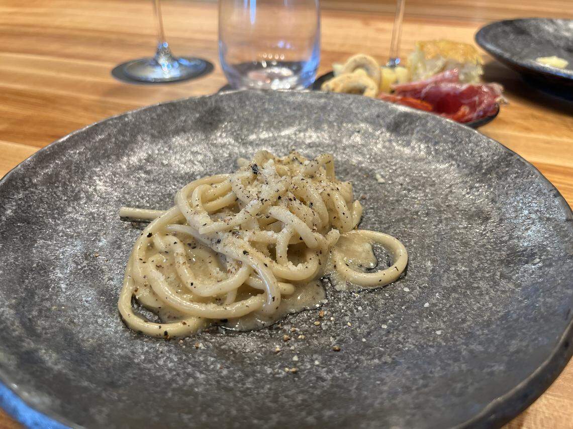A high-angle shot of a small serving of cacio e pepe pasta on a dark, textured gray plate. The thick spaghetti or bucatini noodles are coated in a creamy, light-colored sauce and heavily seasoned with freshly cracked black pepper. The plate rests on a wooden table, and in the blurred background, other dishes and glassware, including what appears to be a charcuterie board with cured meat, are visible.