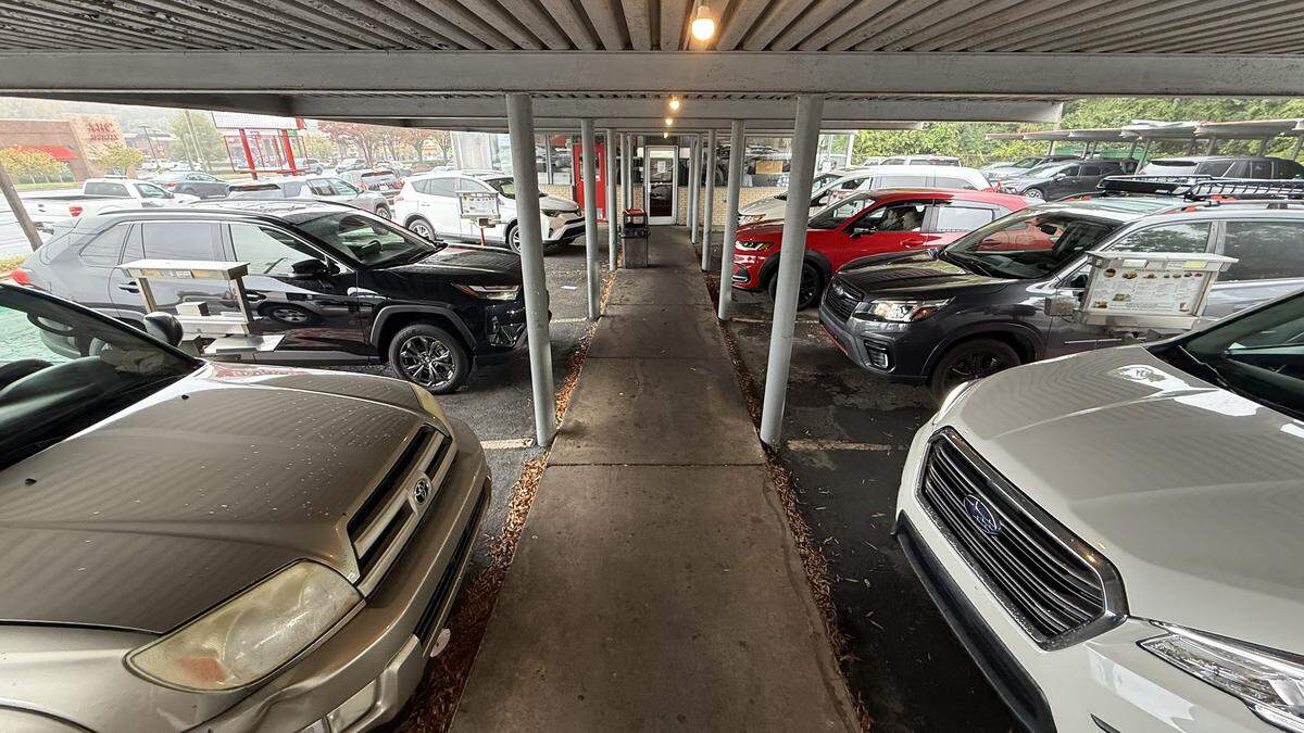 A long, narrow walkway under the covered awning of a drive-in restaurant. Cars are parked at an angle on both the left and right sides of the walkway, facing outward. The restaurant’s building is visible at the far end of the covered path.