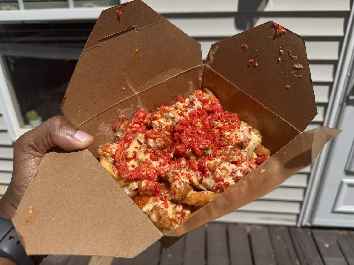 A first-person view of a hand holding an open brown cardboard takeout box filled with golden fries, heavily smothered in creamy white cheese sauce and topped with a generous amount of crushed bright red spicy snacks.
