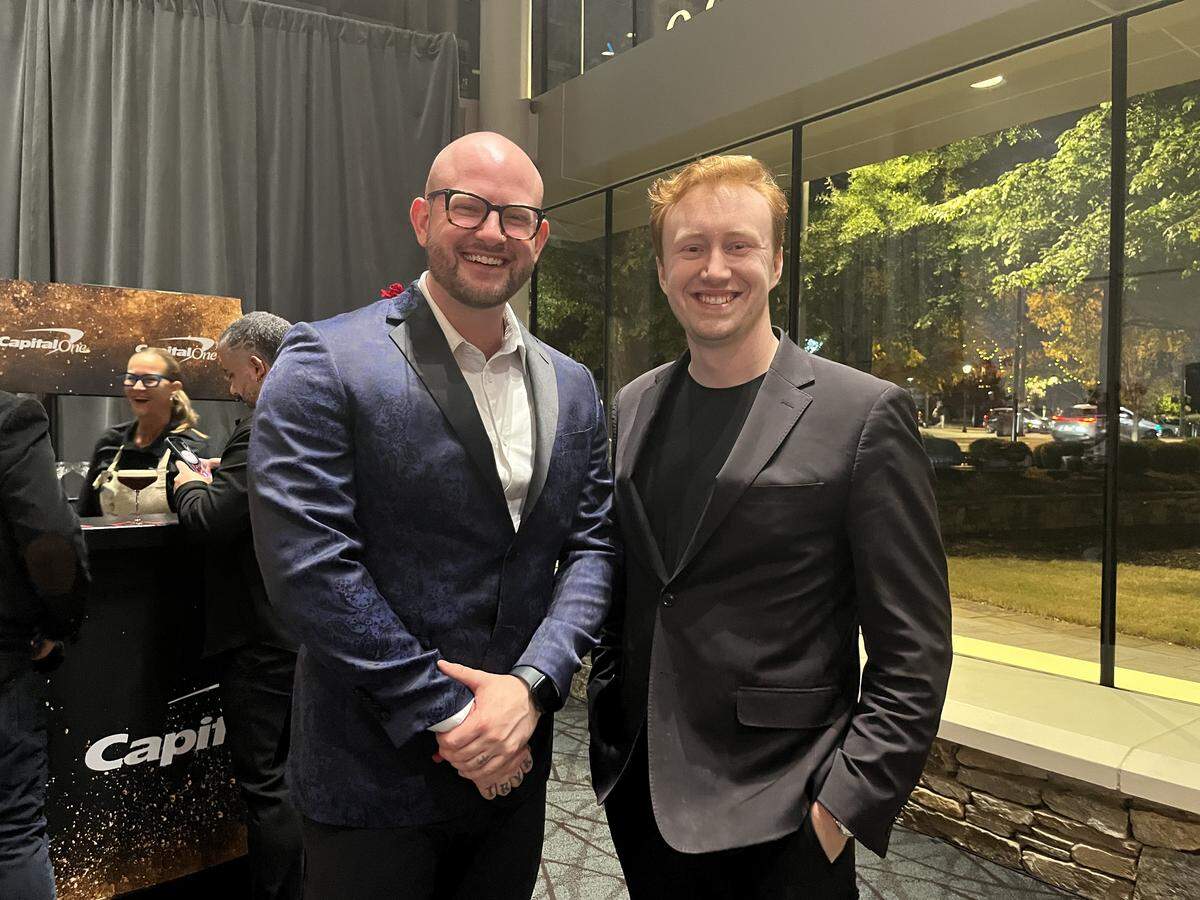 A head-on, medium shot of two people smiling and posing for a photo at an indoor event. The one on the left is bald with glasses, wearing a blue patterned blazer, and the one on the right has red hair and is wearing a black blazer with a black shirt. In the background, a “Capital One” branded bar is visible to the left, and a large window to the right looks out on an illuminated outdoor area.