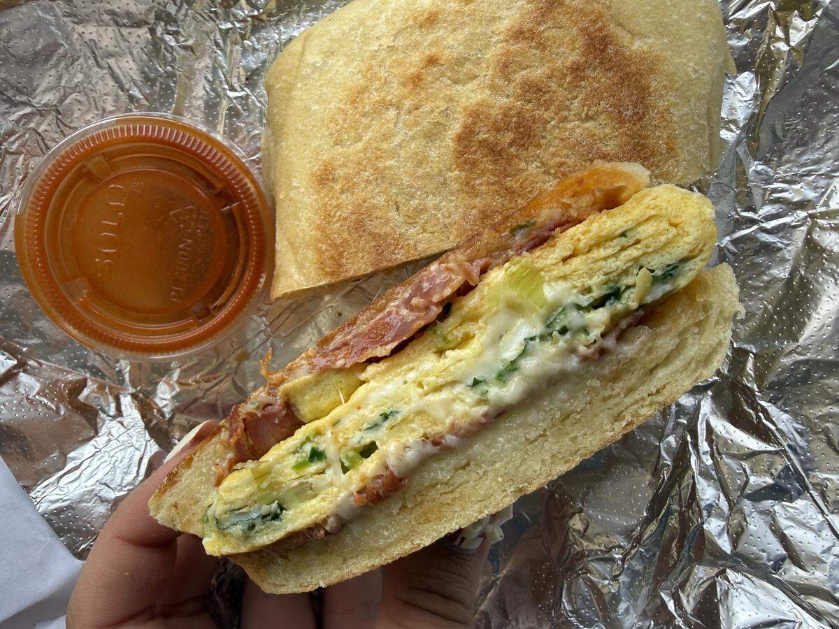 A close-up, overhead shot of a person holding half of a breakfast sandwich on pressed bread. The sandwich is cut to reveal layers of folded egg mixed with green onions, melted white cheese, and crispy chorizo. The sandwich rests on crinkled aluminum foil, with the other half and a small plastic cup of red sauce visible.
