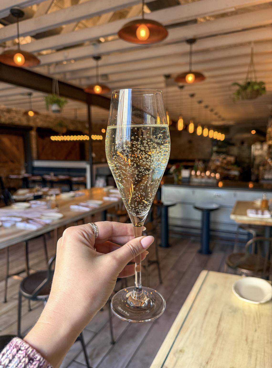 A close-up of a hand holding a champagne flute filled with sparkling wine, raised in a toast on the rustic, candle-lit outdoor patio of Haberdish restaurant in Charlotte, NC.