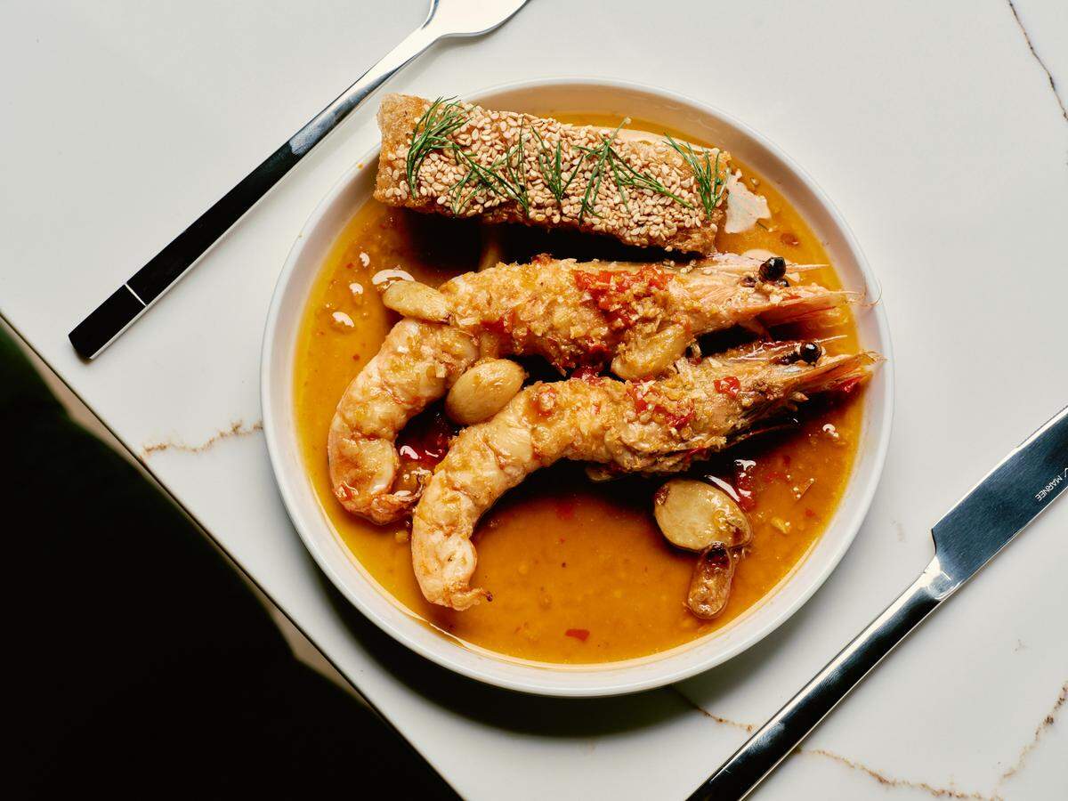 A close-up, top-down view of a gourmet seafood dish in a white bowl set on a marble surface. The bowl contains three large prawns and a rectangular, sesame-seed-crusted piece of fish resting in a golden-colored broth with roasted garlic cloves. A fork and knife are placed on either side of the bowl.