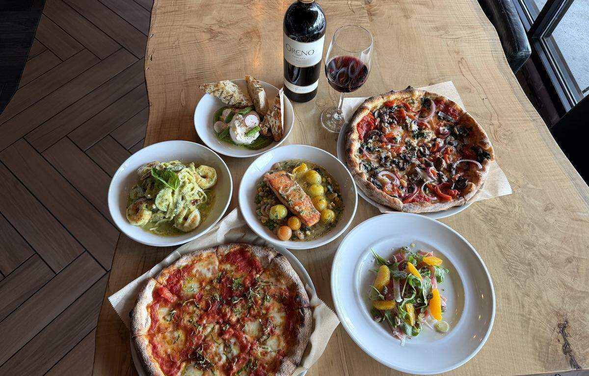 A flat-lay view of an Italian feast on a light wood table. The spread includes two wood-fired pizzas, a bowl of shrimp pesto pasta, a seared salmon fillet with yellow cherry tomatoes, a burrata appetizer with crostini, and a fresh citrus salad. A bottle of red wine and a filled wine glass are centered at the top of the arrangement.