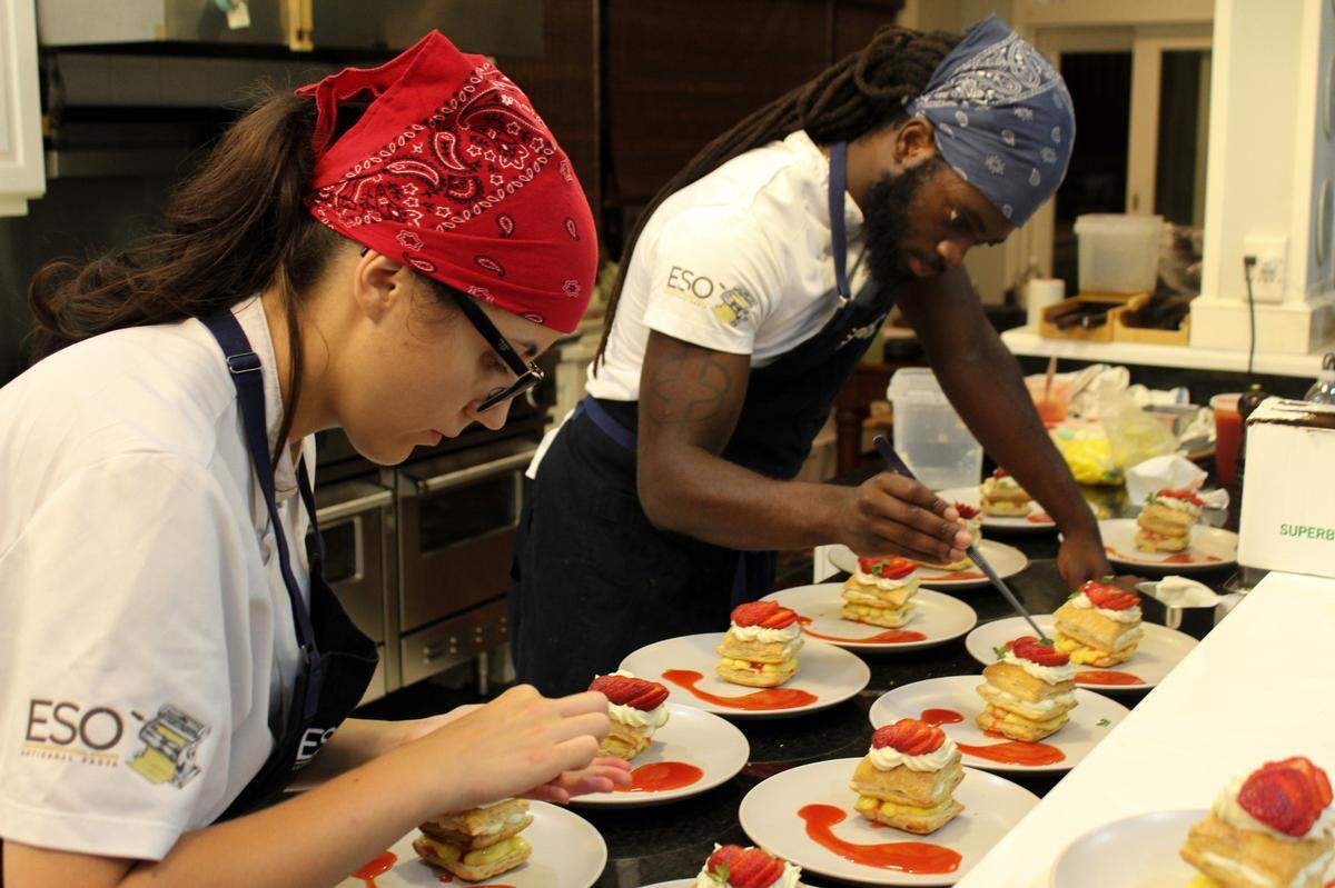 Two chefs in a professional kitchen meticulously plating desserts. On the left, one chef in a red bandana and glasses carefully arranges a pastry, while on the right, a chef in a blue bandana uses tweezers to garnish identical multi-layered desserts topped with fresh strawberries and a red sauce drizzle. Both wear white “ESO” branded shirts and dark aprons.