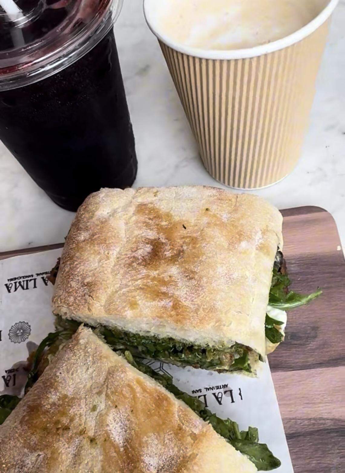 An overhead view of a meal on a white table. A sandwich is cut in half on a wooden board with branded paper. Next to it is a dark iced beverage in a clear plastic cup and a hot latte in a tan paper cup.