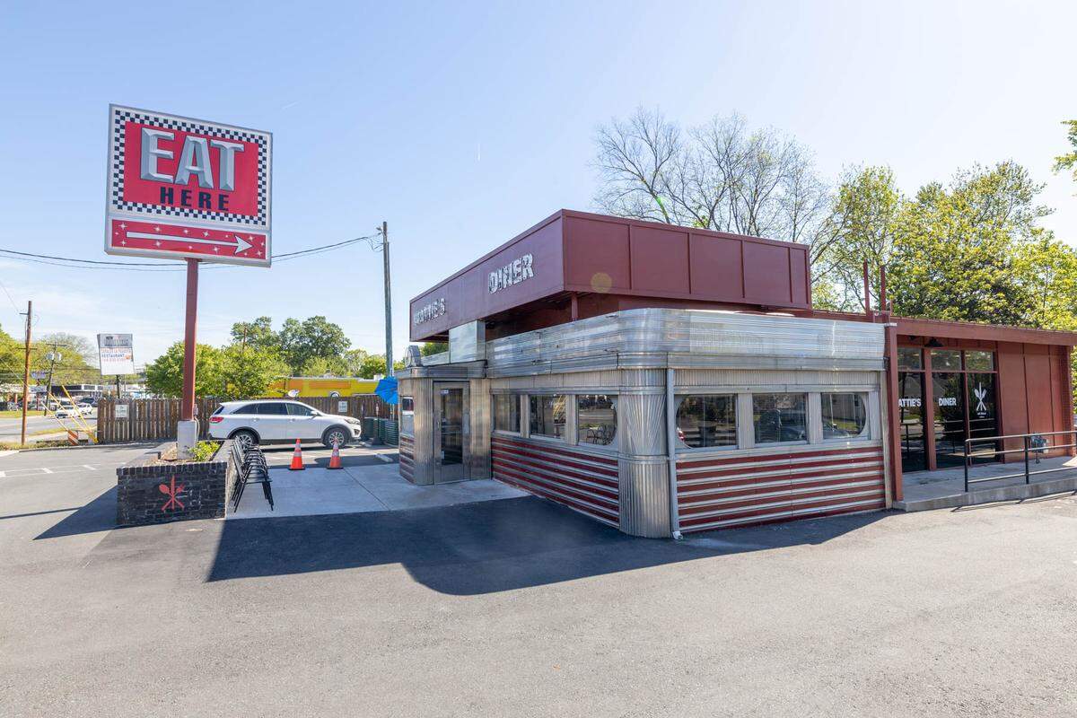 A full, exterior view of a diner named Mattie’s Diner. The building has a classic, stainless-steel diner car section with red stripes, connected to a modern section with large windows and a reddish-brown facade. A retro sign on a pole with an arrow pointing to the right reads “EAT HERE.” A white SUV is parked in the background. The sky is bright blue and there are trees with green leaves in the background.