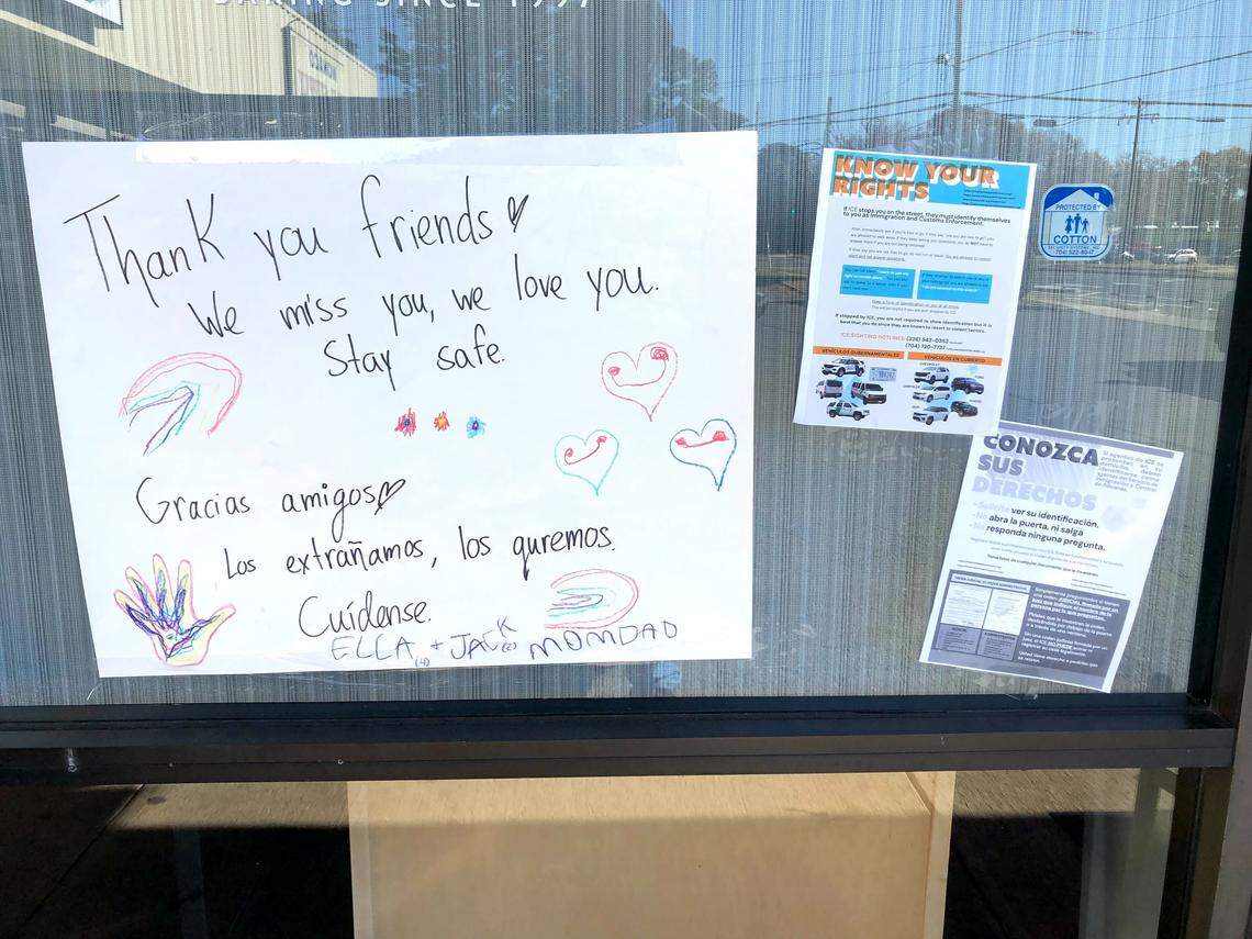 A handwritten note taped to a glass storefront window, surrounded by other notices. The main note, decorated with crayon drawings of hearts, a rainbow, and a handprint, reads: “Thank you friends! We miss you, we love you. Stay safe. Gracias amigos. Los extrañamos, los queremos. Cuídense. ELLA + JACK + MOMDAD.” Other signs include one in English titled “KNOW YOUR RIGHTS” and its Spanish counterpart, “CONOZCA SUS DERECHOS.”