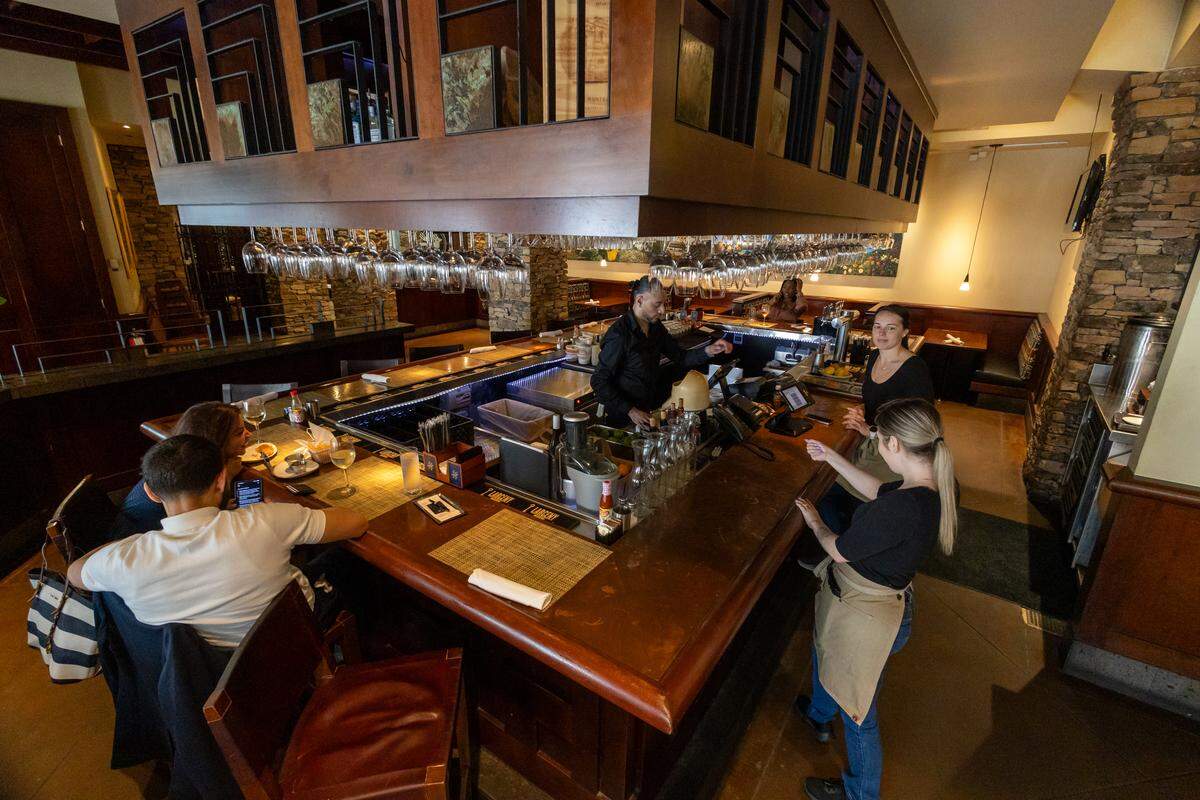 An interior view of the restaurant’s dark wood bar. A bartender in a black shirt is working behind the counter while two servers in black shirts and tan aprons stand nearby. A large, square wooden structure hangs above the bar, holding rows of inverted wine glasses.