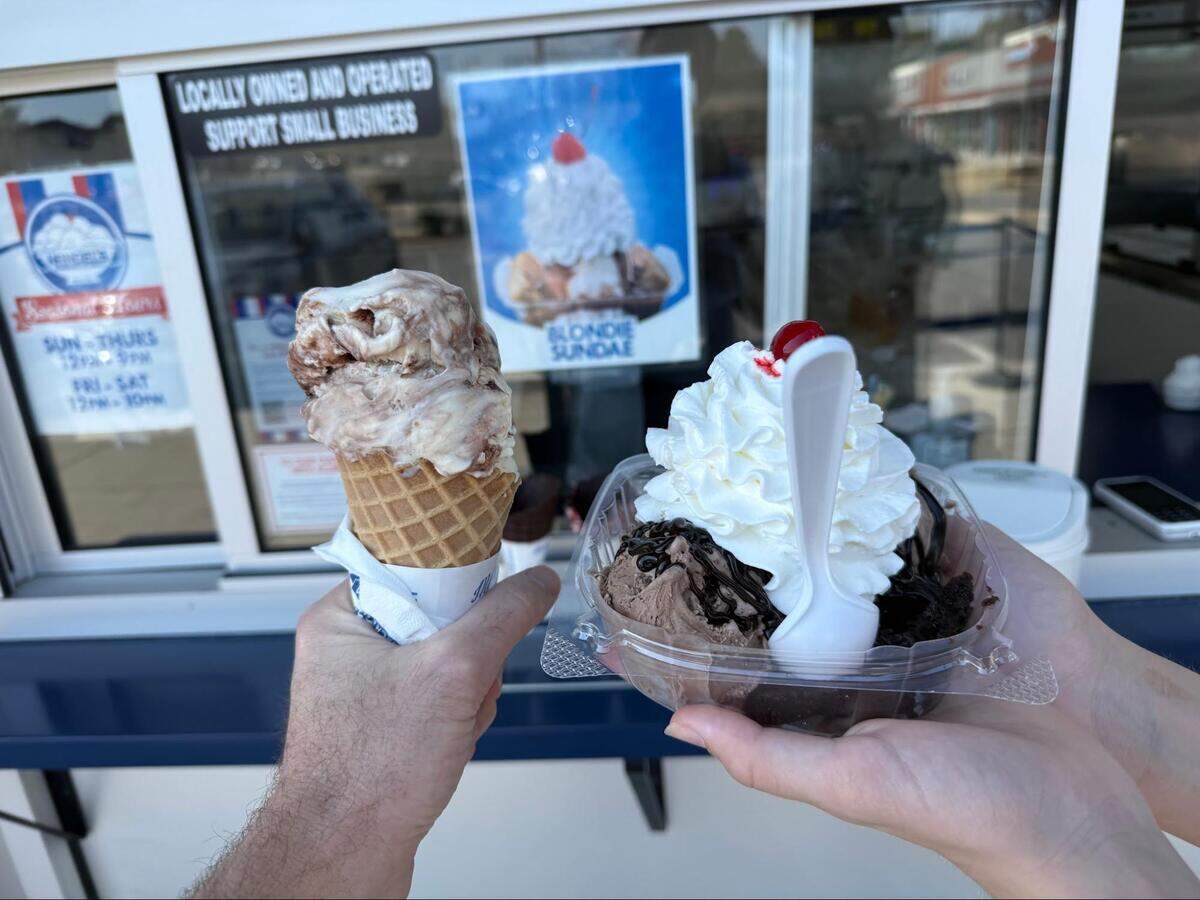 A close-up of two hands holding a marbled ice cream waffle cone and a chocolate sundae topped with whipped cream and a cherry.