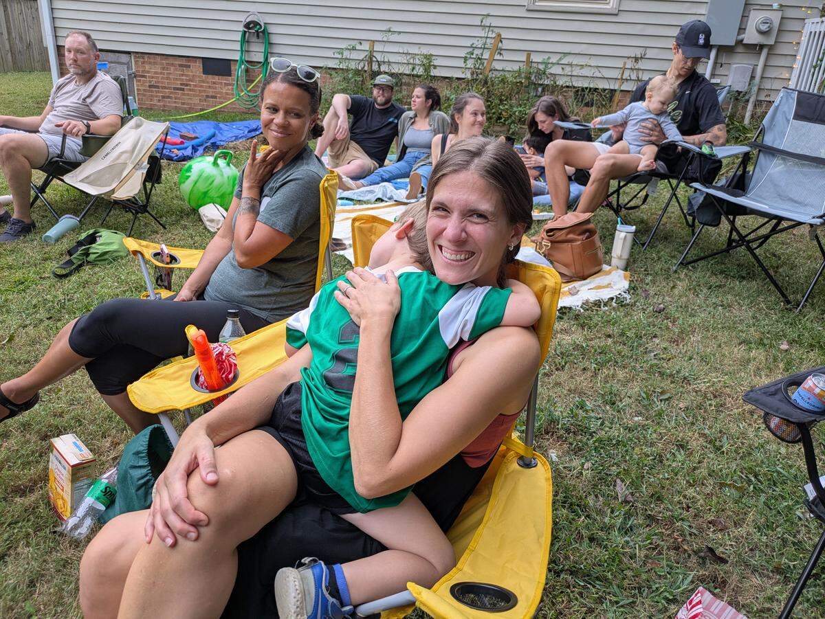 A close-up, high-angle shot of a mother smiling at the camera while sitting in a yellow camping chair and holding a small child amid a larger group of adults and children relaxing in a grassy backyard, seated on blankets and in other lawn chairs, enjoying a casual outdoor gathering.