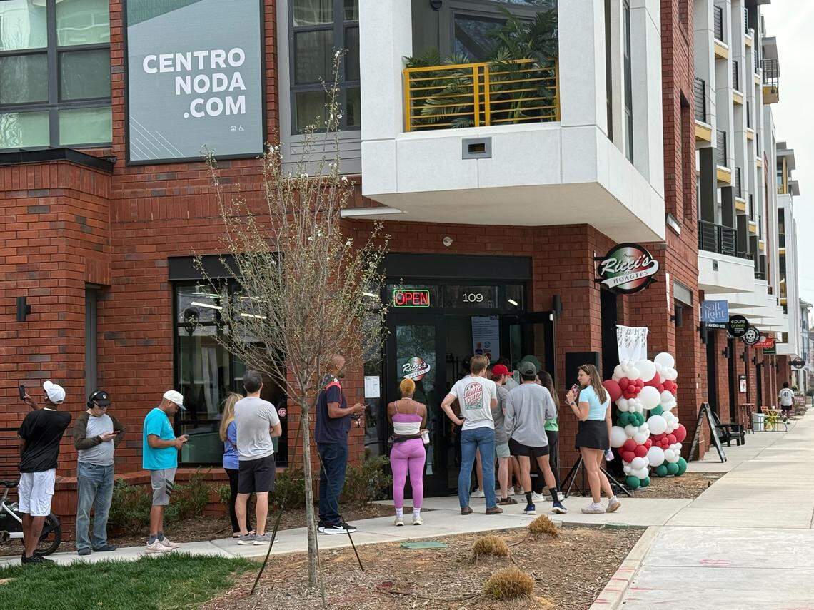 A close-up view of the entrance to “Ricci’s Hoagies,” where a group of people is gathered near the door. An “Open” sign is lit in the window, and a decorative arch of red, white, and green balloons stands next to the entrance. Several people in line are looking at their phones while waiting.