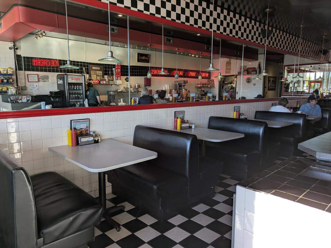 A wide shot of the interior of a classic 1950s-style diner. The foreground shows black vinyl booths and white-topped tables on a black and white checkered floor. A long red counter separates the dining area from the service area, which has white tiled walls and a large mirror reflecting the restaurant. Several patrons are seated at the counter and in the booths, and a red digital sign reading “ICE CREAM” (backwards) hangs in the back.
