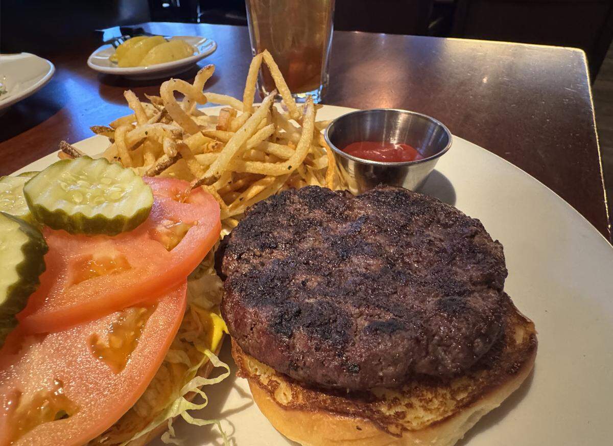 A close-up, high-angle shot of a gourmet burger meal served on a white ceramic plate. The meal features a thick, seared beef patty on a toasted bun, accompanied by a large pile of thin, golden-brown shoestring fries and a small metal ramekin of ketchup. The open-faced side of the bun is layered with shredded lettuce, two large slices of tomato, and several crinkle-cut pickles.