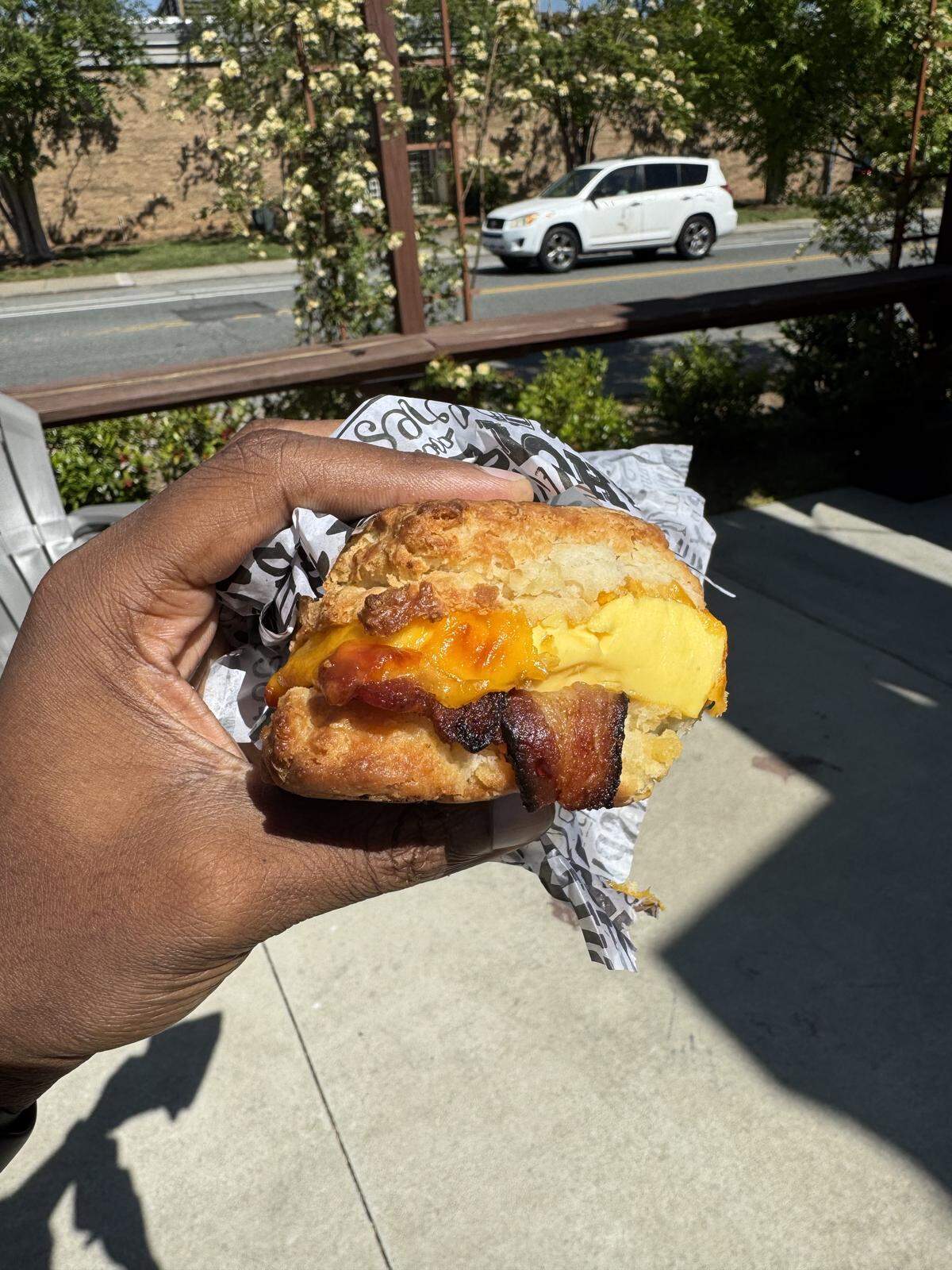 A close-up shot of a breakfast biscuit held in a hand outdoors. The biscuit is filled with a folded yellow egg, melted cheddar cheese, and crispy strips of bacon. The scene is brightly lit by sunlight, with a sidewalk and a white car visible in the blurred background.