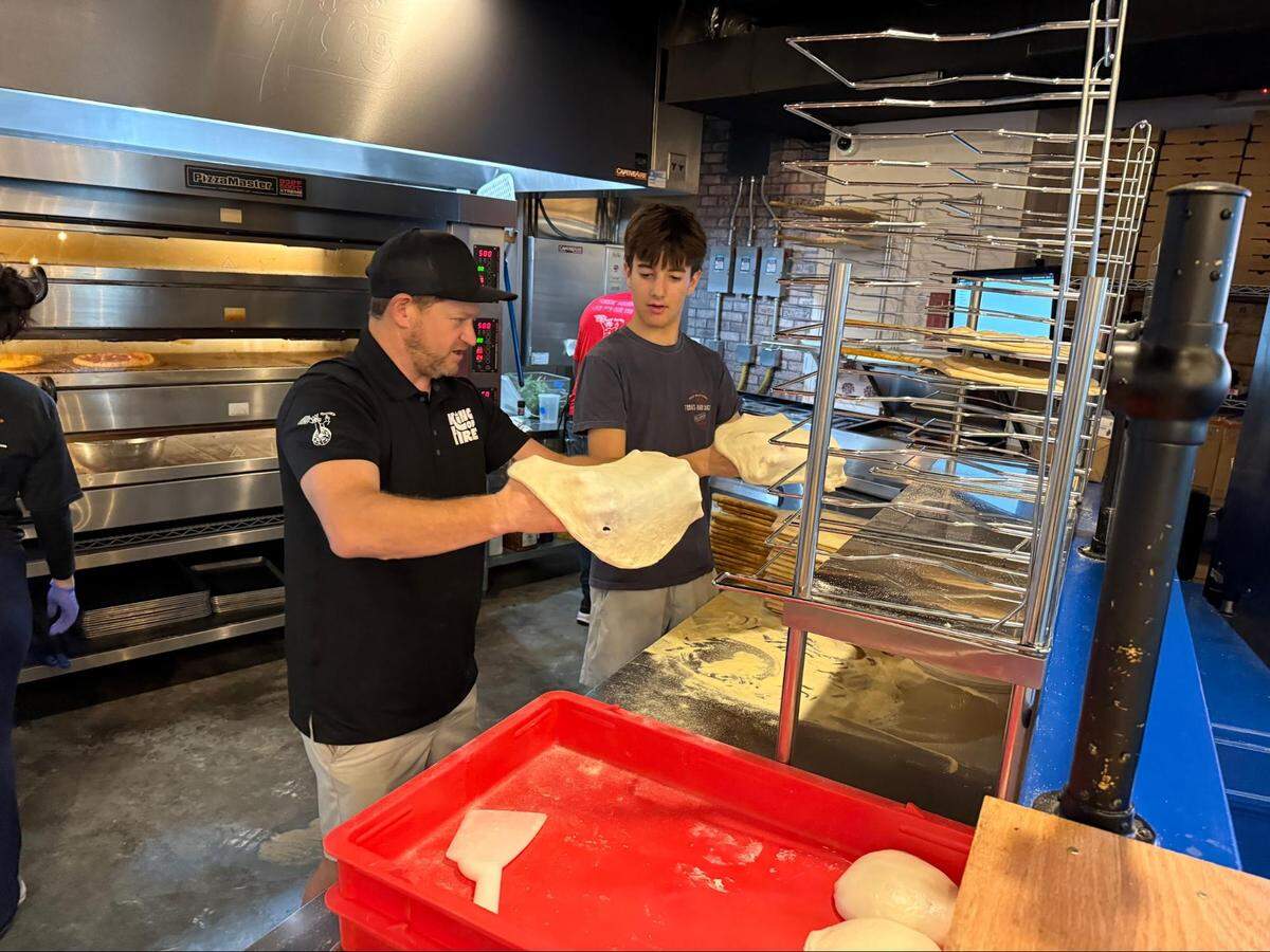 In a commercial kitchen, a chef in a black “King of Fire” uniform demonstrates how to stretch pizza dough. A young person stands by holding another piece of dough and watching the technique closely. A red bin containing fresh dough balls is in the foreground.
