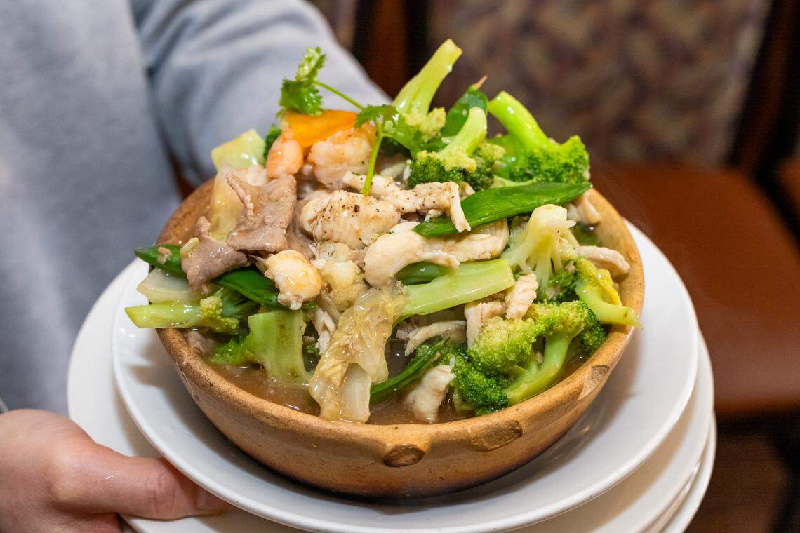 A close-up shot of a person holding a large, rustic wooden bowl filled with a stir-fry. The dish is overflowing with shrimp, chicken, and other meats, mixed with vibrant green broccoli, snow peas, and cabbage in a light brown sauce, and garnished with a sprig of cilantro. The bowl is set on a white plate.