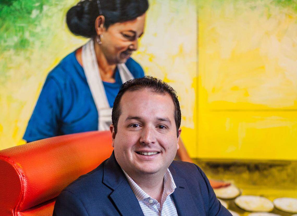 A professional headshot of a business owner with dark hair smiling warmly at the camera and wearing a navy blazer over a collared shirt. The person is seated in a bright red booth, and in the background is a large, colorful painting with a yellow and green background that depicts a woman preparing food.
