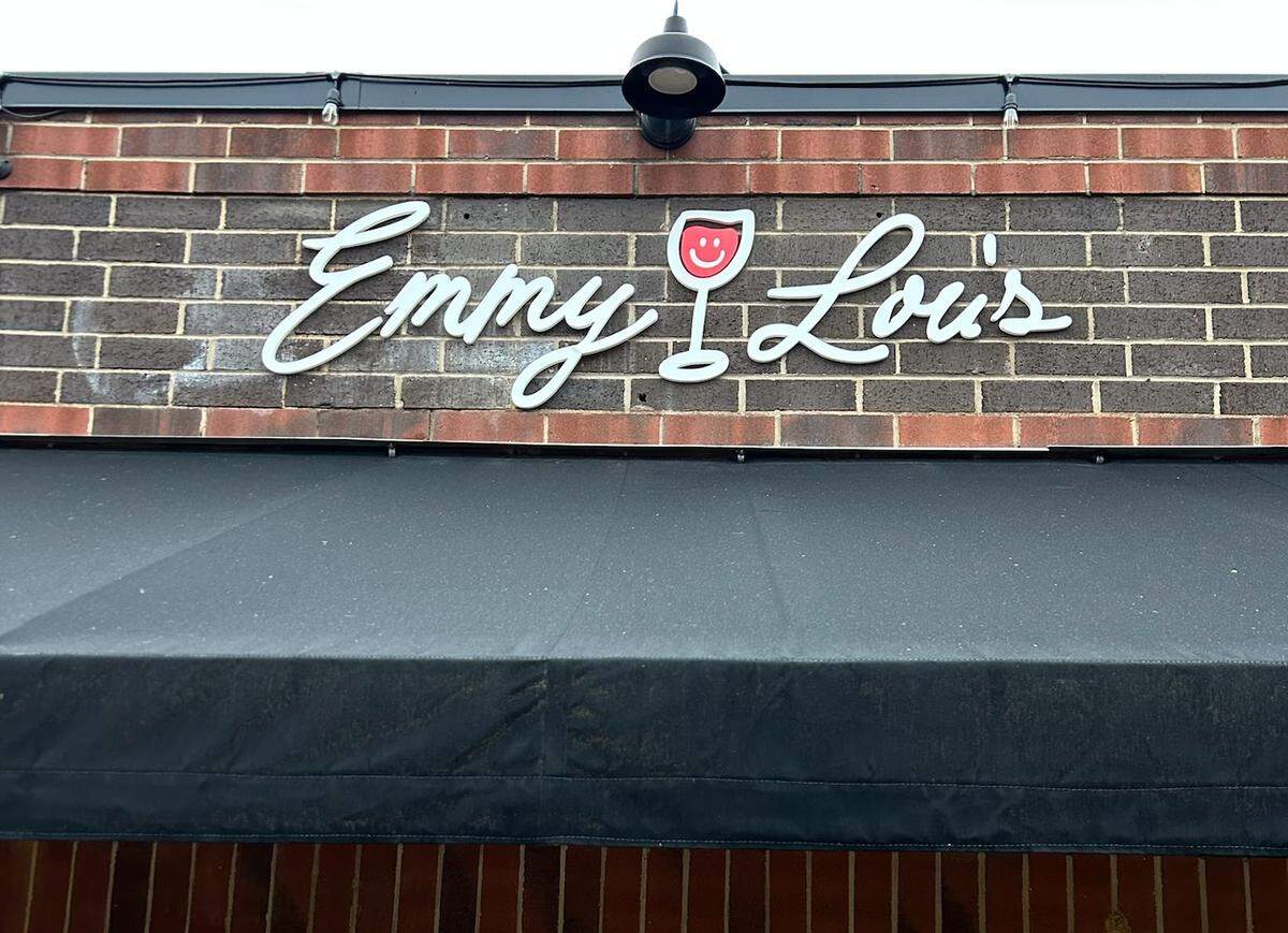 A close-up of the storefront sign for a business named ‘Emmy Lou’s.’ The name is written in white cursive letters on a dark red brick wall, with the ‘o’ in ‘Lou’s’ replaced by a graphic of a wine glass containing a red smiley face. A black awning stretches across the bottom of the frame, and a single black industrial light fixture is mounted above the sign