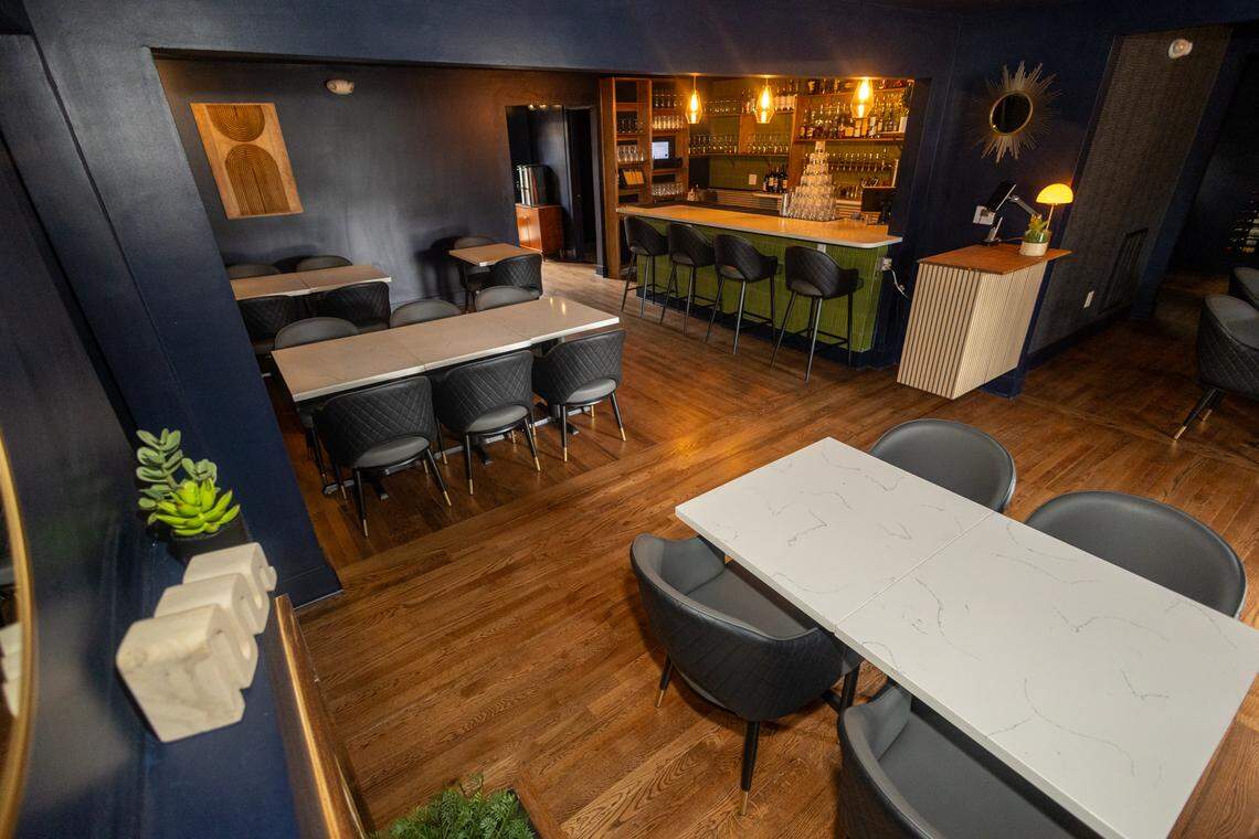 A stylish restaurant and bar interior with dark blue walls and a warm, inviting atmosphere. The room has light wood flooring and is furnished with several tables featuring white faux marble tops and dark gray chairs. In the back, a bar area is illuminated by warm lighting, with a green bar counter and black barstools. The decor includes modern artwork, a sunburst mirror on the wall, and various potted plants. The lighting is a mix of recessed spotlights and hanging fixtures, casting a cozy glow on the space.