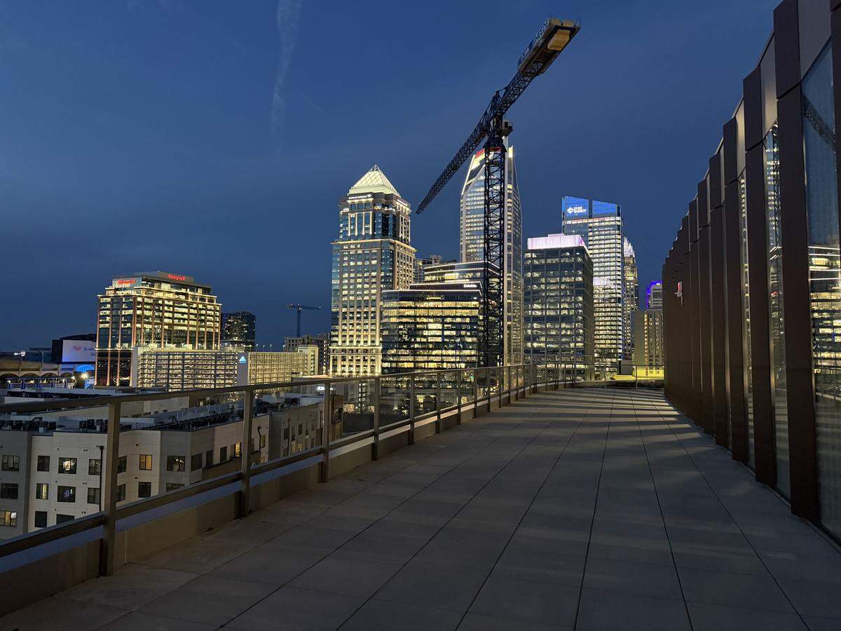 A wide-angle landscape photograph captures the expansive outdoor rooftop patio of a building in Charlotte, North Carolina, during twilight. Visible landmarks include the Truist Center with its distinctive curved, glowing roof and several other high-rise office buildings with various lighting displays. A large construction crane stands prominently in the center of the skyline, indicating ongoing urban development.