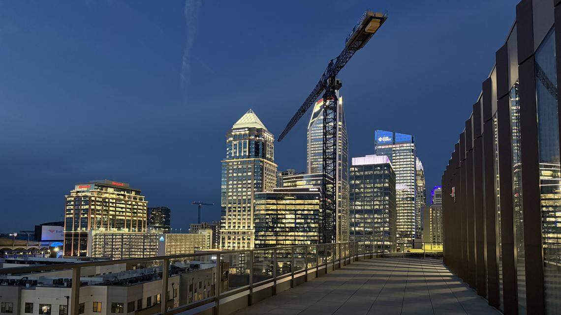 A wide-angle landscape photograph captures the expansive outdoor rooftop patio of a building in Charlotte, North Carolina, during twilight. Visible landmarks include the Truist Center with its distinctive curved, glowing roof and several other high-rise office buildings with various lighting displays. A large construction crane stands prominently in the center of the skyline, indicating ongoing urban development.