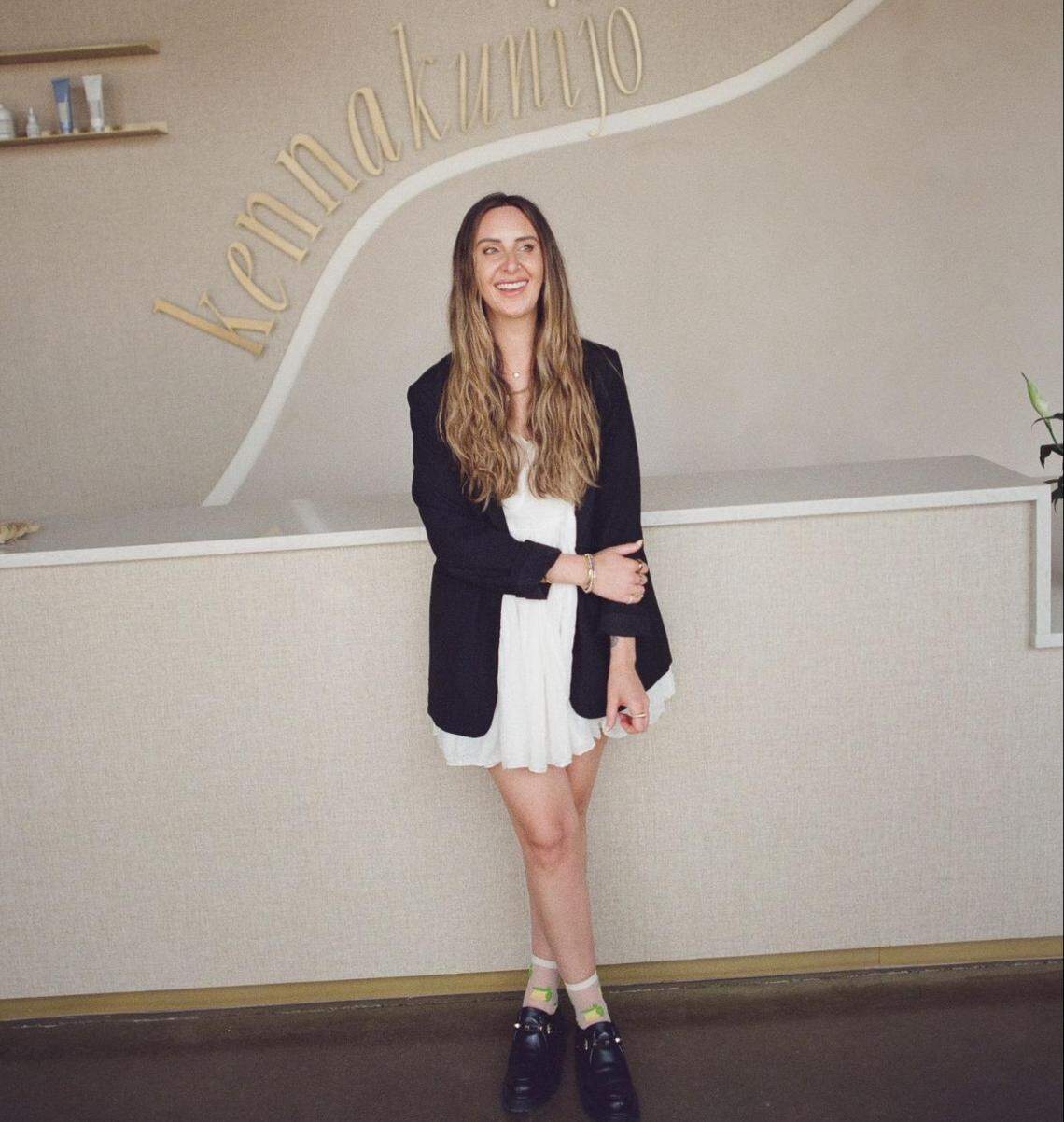 A person with long blonde hair smiles while standing in front of a modern reception desk. The wall behind the person features the gold-colored name “Kennakunijo” in a curved font.