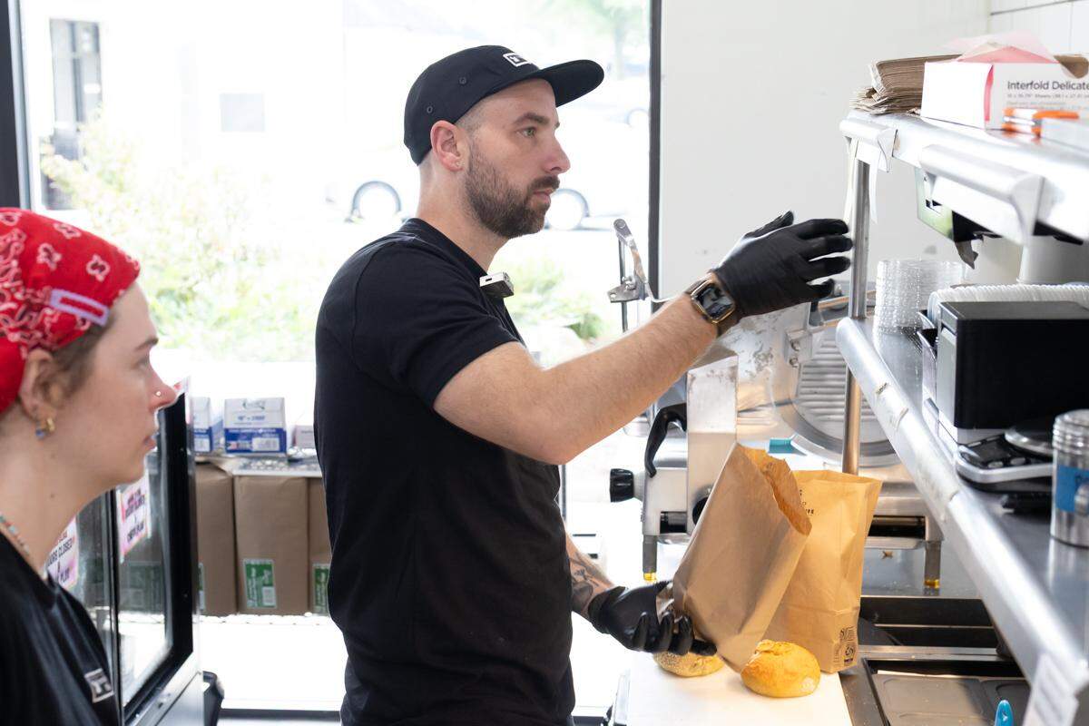A food service worker wearing a black t-shirt, a black baseball cap, and black gloves is reaching for a container on a metal shelf behind a counter. Another worker, wearing a red bandana, is visible in the foreground on the left, looking in the same direction. They are in a well-lit commercial kitchen or bakery.