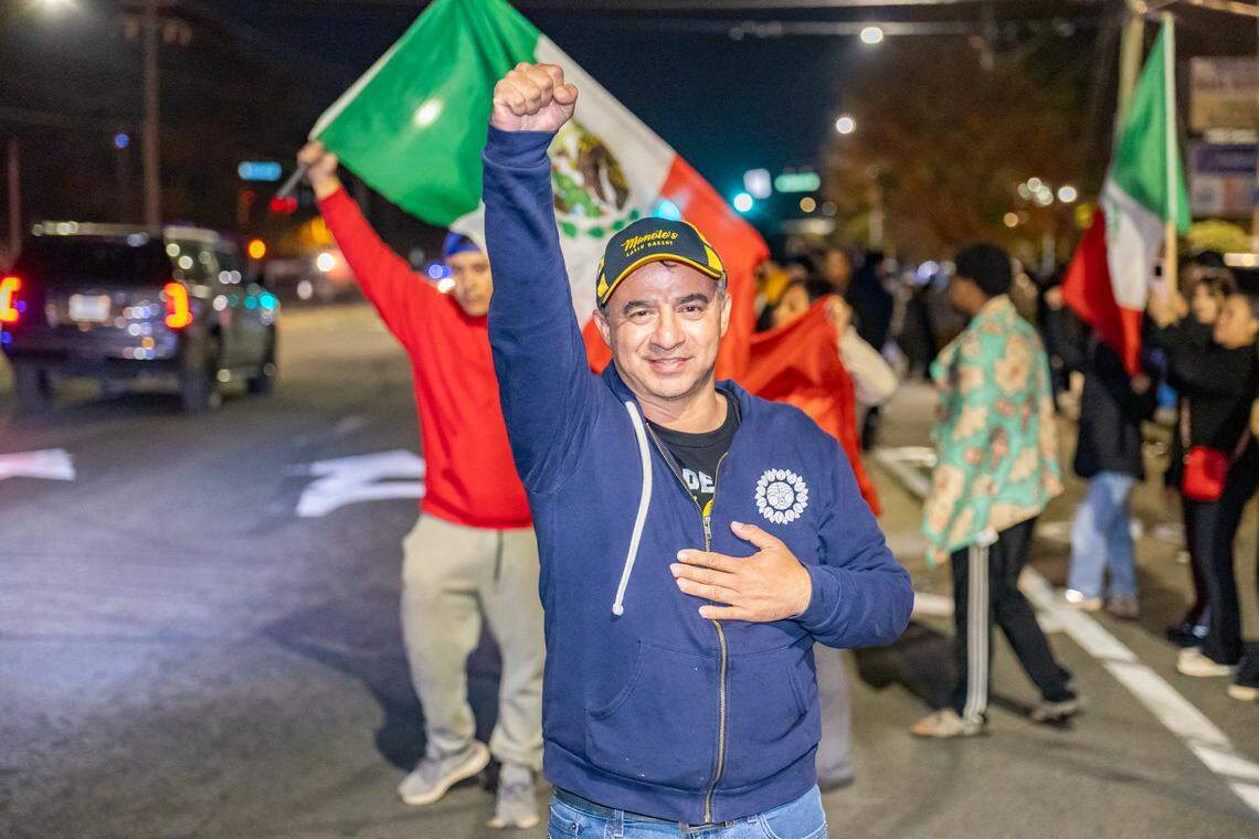 Bakery owner Manuel “Manolo” Betancur, wearing a blue jacket and baseball cap, walks across a dark paved area at night, raising one hand. In the background, a large crowd of protesters is visible on the street and sidewalk.