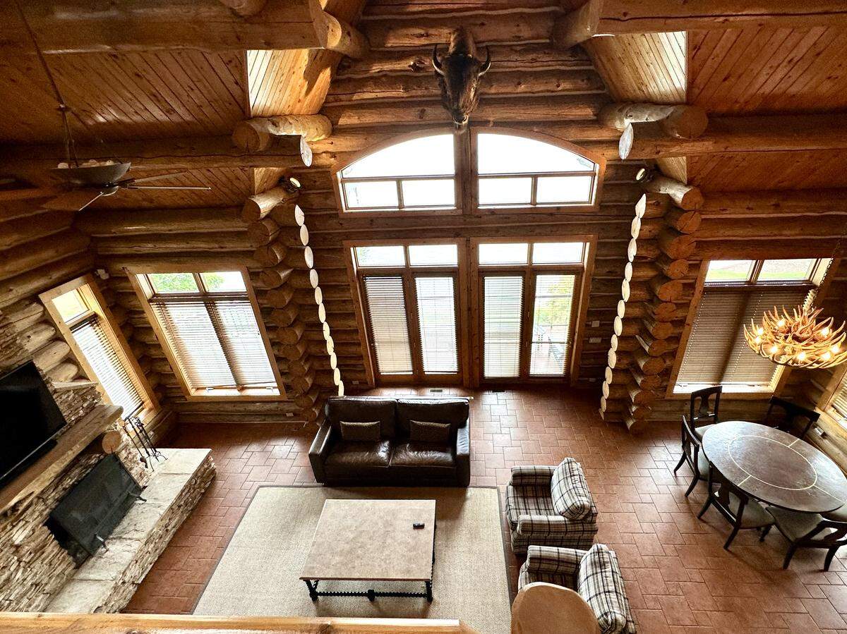 A high-angle view looking down into the great room of a large log cabin. The room features a vaulted ceiling with exposed log beams, a stone fireplace, and a dining area with an antler chandelier. The living space is furnished with a leather sofa, two plaid armchairs, and a coffee table.