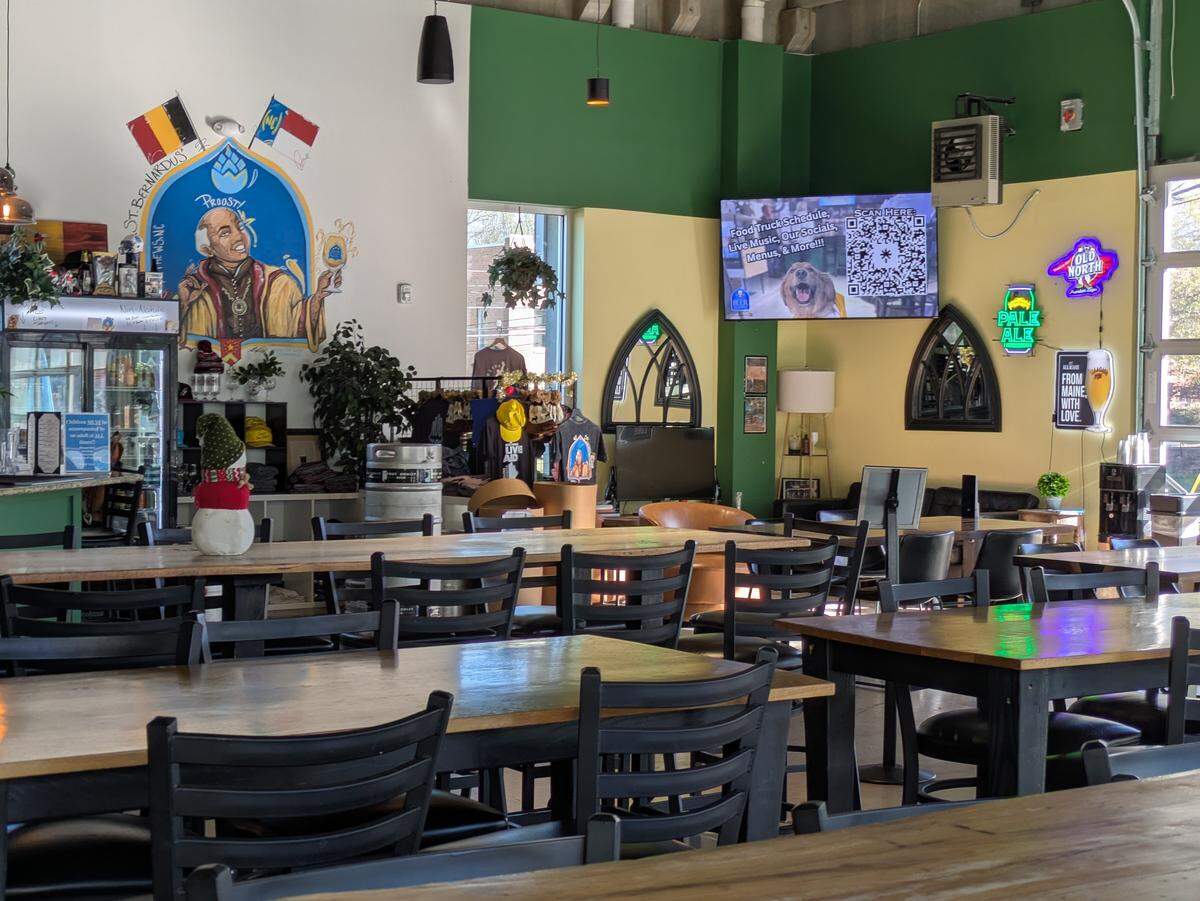 The interior seating area of a brewery with long wooden communal tables and black chairs. The walls are painted green and yellow, featuring a large colorful mural of a monk holding a beer glass under the words “St. Bernardus” and “Proost!”