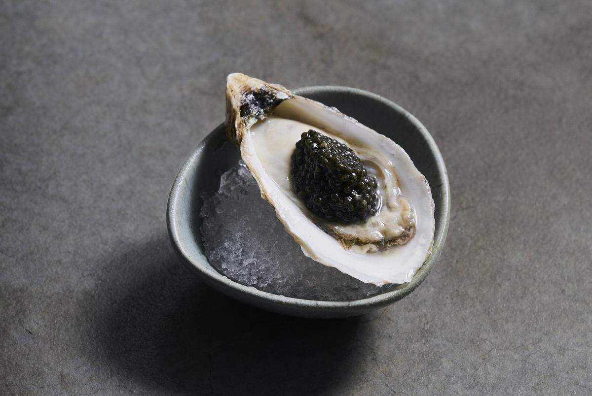 high-angle, close-up shot of a single fresh oyster served on the half shell, presented as a gourmet delicacy. The oyster rests at a slight angle on a bed of finely crushed ice inside a small, rustic, organic-shaped ceramic bowl with a matte, sage green finish. Nestled perfectly inside the glistening oyster shell, a generous spoonful of glossy, dark green-to-black caviar serves as the centerpiece.