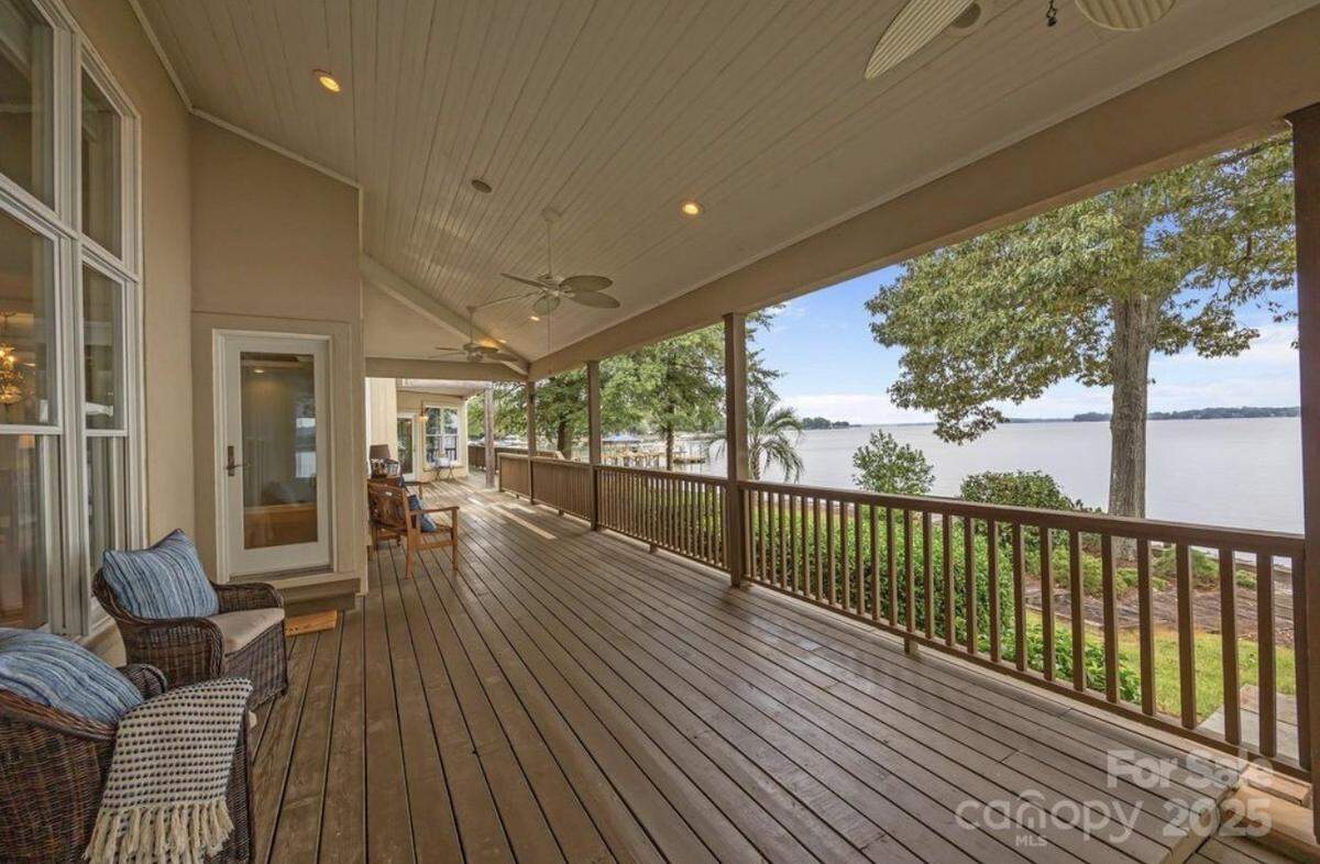 A wide-angle view of a deep, covered wooden deck or porch that runs along the back of a house, featuring ceiling fans, recessed lighting, and a few wicker chairs with blue pillows. The deck is enclosed by a dark wood railing and offers a stunning, unobstructed, panoramic view of a large lake framed by mature trees.