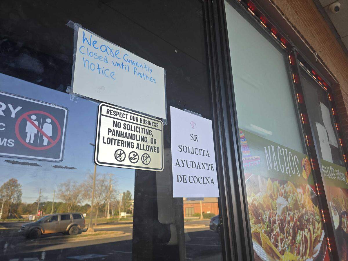 A close-up of a glass storefront window showing several signs. A handwritten note in blue marker on a white sheet of paper reads: “We are currently closed until further notice.” Below it is a black and white sign with a crossed-out symbol of a person, and a text box that reads: “RESPECT OUR BUSINESS NO SOLICITING, PANHANDLING, OR LOITERING ALLOWED.” To the right is a white sign in Spanish that reads: “SE SOLICITA AYUDANTE DE COCINA” (Kitchen helper wanted). The reflection of a parked vehicle and surrounding area is visible in the glass.