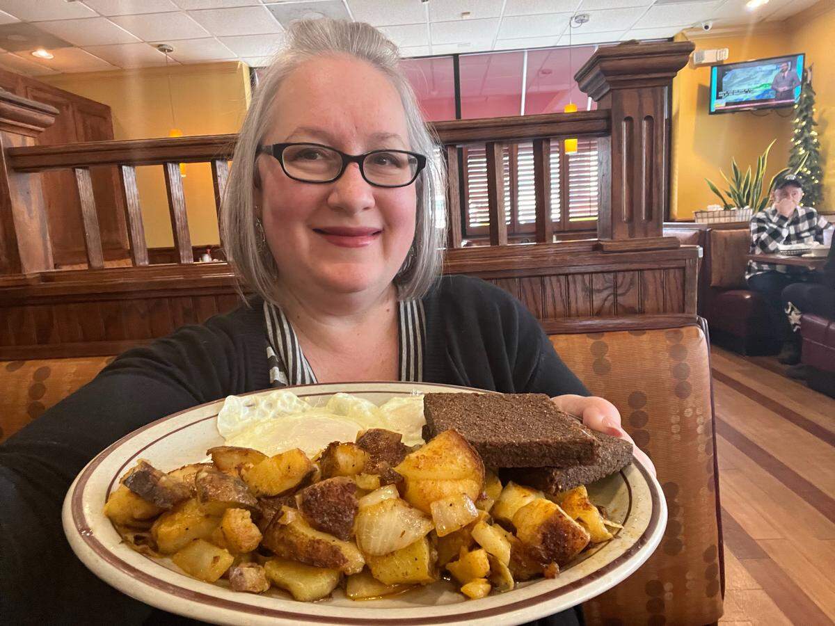 A restaurant customer with glasses smiles while holding up a large plate of breakfast food in a restaurant booth. The plate contains home fried potatoes and onions, two fried eggs, and two slices of livermush.