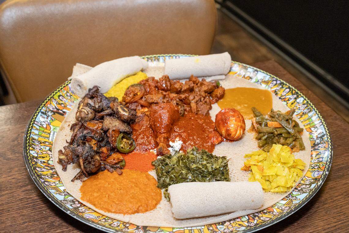 A top-down view of a colorful and diverse Ethiopian feast. The large silver tray is lined with injera and topped with a variety of traditional dishes, including Doro Wot (spicy chicken stew with a hard-boiled egg), Gomen (collard greens), and various lentil and vegetable stews. Several rolls of extra injera are placed around the edges, ready for guests to use as utensils for the communal meal.
