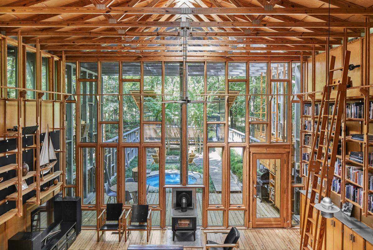 A wide, symmetrical shot of the interior of a modern, wood-framed cabin. The entire back wall is a floor-to-ceiling grid of windows that looks out onto a wooden deck, a small blue circular pool, and a dense forest. The room has a high, vaulted ceiling with exposed wooden trusses. The interior is flanked by a large, built-in bookshelf with a rolling ladder on the right and another wooden shelving unit on the left. A black wood-burning stove sits in the center of the room.