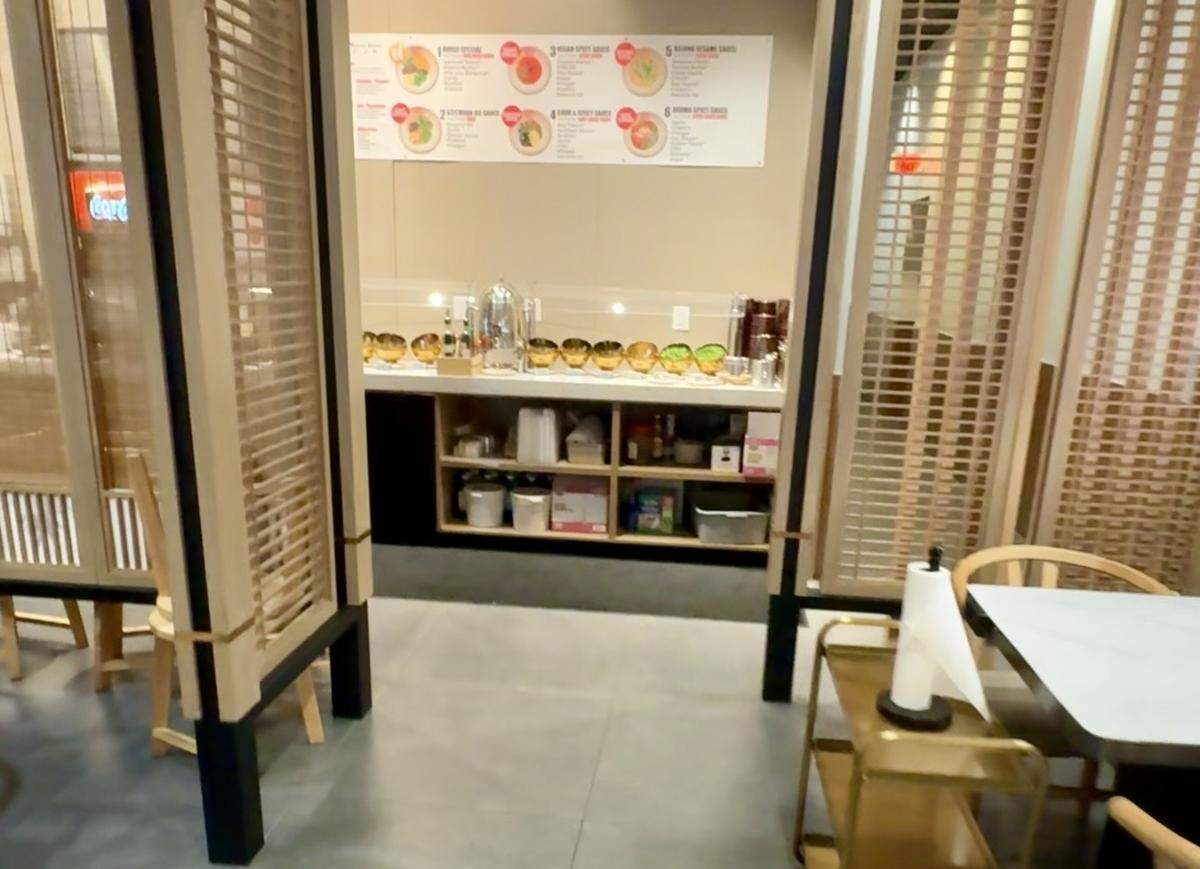 A wide shot of a self-serve sauce station in a restaurant. A long white counter holds various golden bowls and dispensers containing condiments and garnishes. Above the counter, a large instructional sign displays six different sauce recipes with photos. The area is framed by modern light-wood partitioned screens.