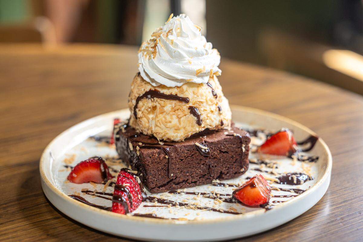 A close-up, appetizing shot of a gourmet dessert. A thick, fudgy chocolate brownie is topped with a large scoop of vanilla ice cream encrusted in toasted coconut and finished with a high swirl of whipped cream. The plate is artistically drizzled with chocolate sauce and scattered with fresh strawberry halves and extra toasted coconut.