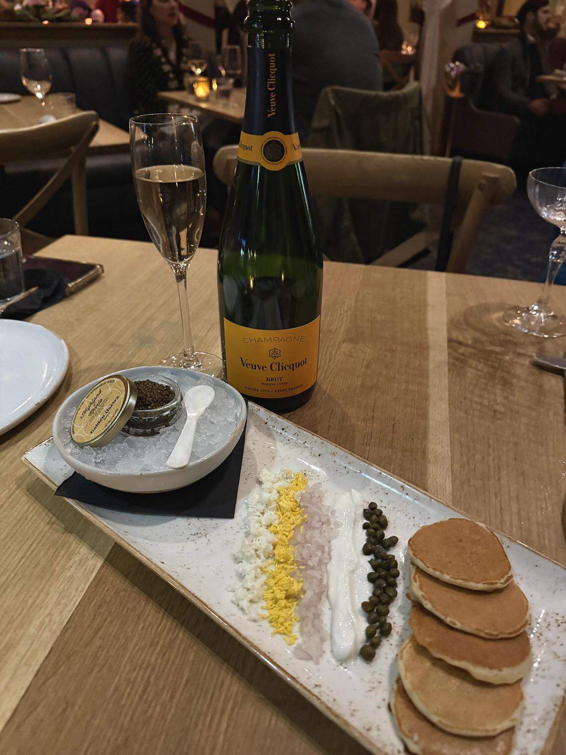 A close-up, slightly high-angle shot of a caviar and champagne service on a light wooden restaurant table. A bottle of Veuve Clicquot Brut Champagne stands next to a tall glass of champagne. In the foreground, a rectangular plate holds small blini pancakes, dollops of sour cream, capers, chopped onions, and egg. A small tin of Golden Osetra caviar sits in a bowl of crushed ice with a white spoon.