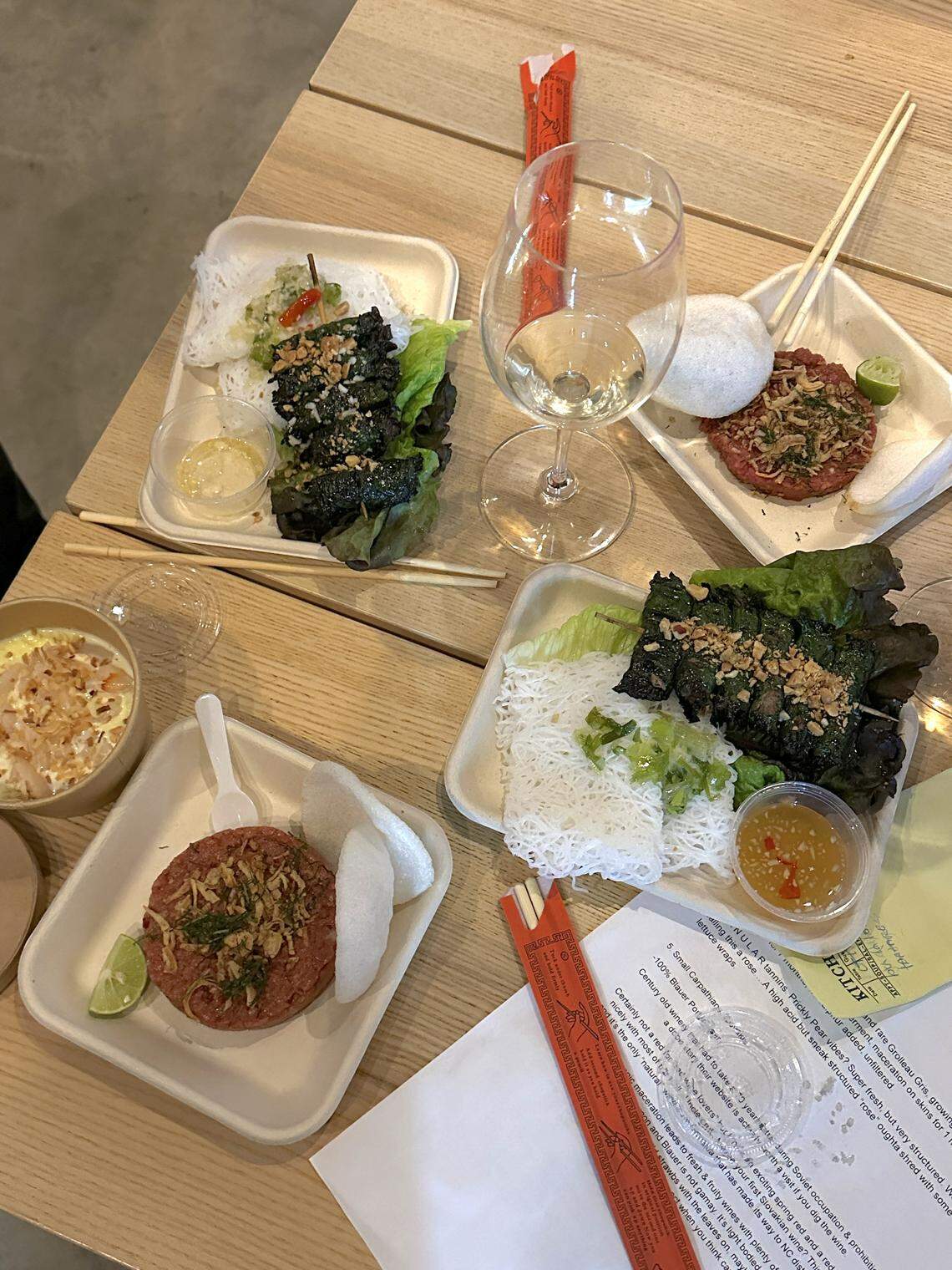 An overhead shot of four Vietnamese dishes arranged on a light wooden table. The dishes include two servings of grilled beef wrapped in betel leaves over vermicelli noodles and lettuce, and two small squares of beef tartare (Bò tái chanh) served with rice crackers and a lime wedge. A wine glass with white wine and two sets of chopsticks are also visible.