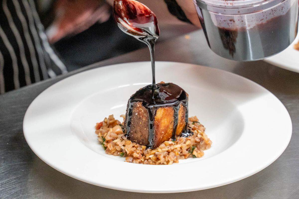 A close-up, action shot of a chef’s hand using a spoon to drizzle a thick, glossy dark balsamic or wine reduction over a square-cut piece of braised meat. The meat sits atop a small bed of textured grains or farro in a wide-rimmed white bowl.