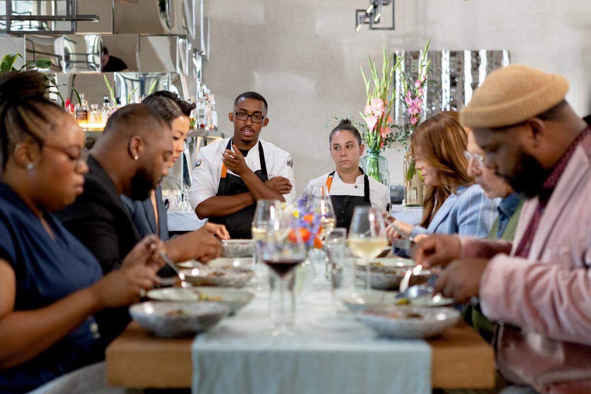An indoor shot of a long dining table where several guests are seated, out of focus in the foreground, eating from shallow bowls. In the background, two chefs in white coats and black aprons stand behind the table. The chef on the left, a man with glasses, is speaking and gesturing with his hand. The chef on the right, a woman with her hair in a bun, listens with a serious expression. The room features modern decor, including a mirrored wall and a tall vase of pink and yellow flowers.