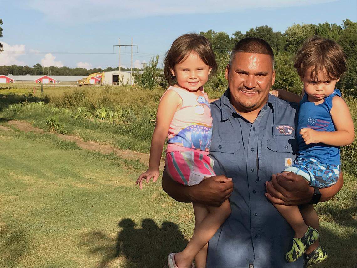 Mountaire chicken farmer Ashley Oxendine holds his two children. Some of his chicken barns are in the background.