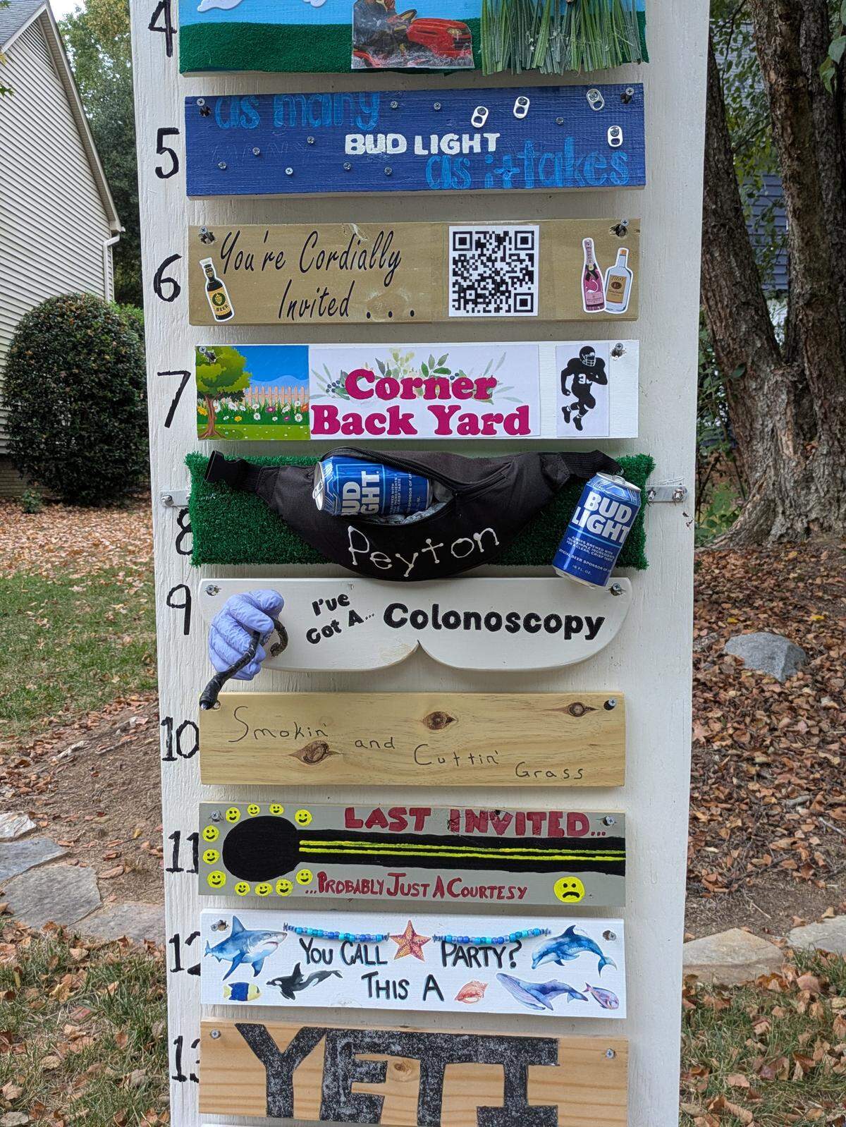 A close-up shot of a tall, white wooden post in a yard, which serves as a humorous, multi-part sign. Numbered planks from 4 to 13 are attached horizontally, each with a different joke or theme. Notable planks include number 8, which has a fanny pack labeled “Peyton” holding a Bud Light can on artificial turf, and number 9, a white cutout plank reading “I’ve Got A... Colonoscopy” with a purple-gloved hand holding a medical tool.