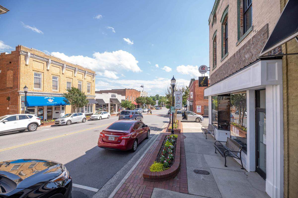 A sunny street view of historic downtown Belmont, North Carolina. Two-story brick buildings line the road, with cars parked along the brick sidewalks. A flower planter and a bench are visible in the foreground under a blue sky.