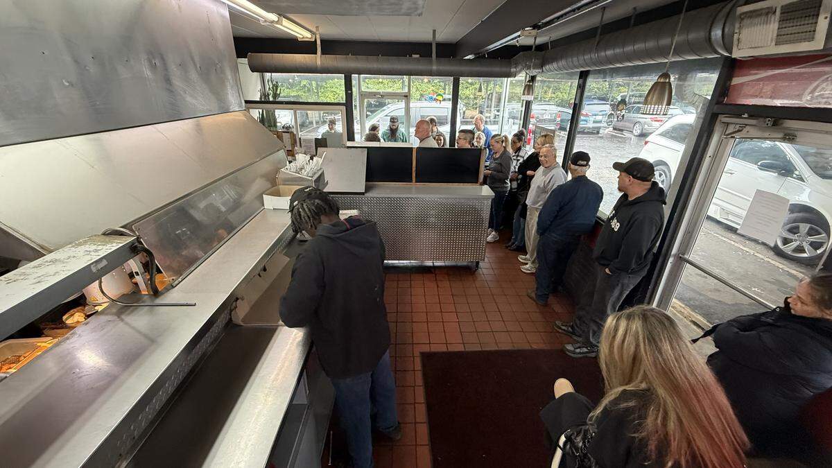 An interior view of a restaurant from behind a stainless-steel service counter. In the foreground, a staff member works at the counter. In the background, a long line of customers is visible, stretching along a wall of windows and out the glass door.