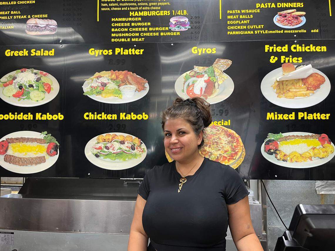 A smiling restaurant owner with dark hair tied back stands in front of a large, black illuminated menu board inside a restaurant. The menu features vibrant photos of various dishes including Greek salads, gyros, kabobs, and fried chicken, with yellow text headings. The owner is wearing a black t-shirt and a gold necklace, positioned centrally in front of the food display.