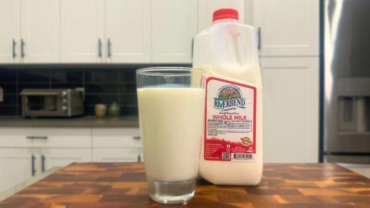 An eye-level shot of a glass of milk next to a gallon jug of Riverbend Creamery Whole Milk, both resting on a wooden butcher block countertop in a modern kitchen with white cabinets.